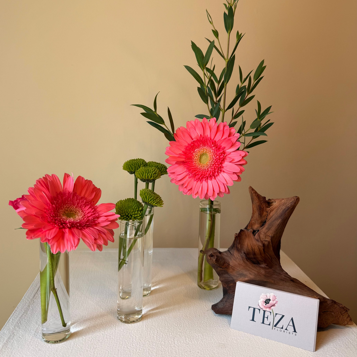 Pink flowers in clear vases on a table with a wooden decorative item and a 'TEZA' sign in the background.